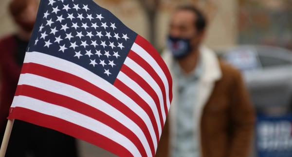 A handheld American flag dominates the foreground of the picture. In the background, we can see the blurry form of a person with short cropped hair, wearing a face mask and a brown jacket over a lighter shirt.